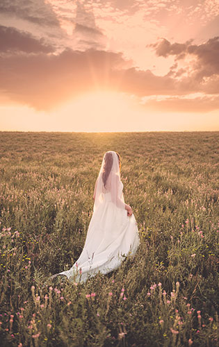 Bride walking alone through a field at sunset on her wedding day.