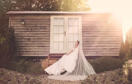 Bride sitting beside a log cabin with her long veil at Wellington Barn in Wiltshire